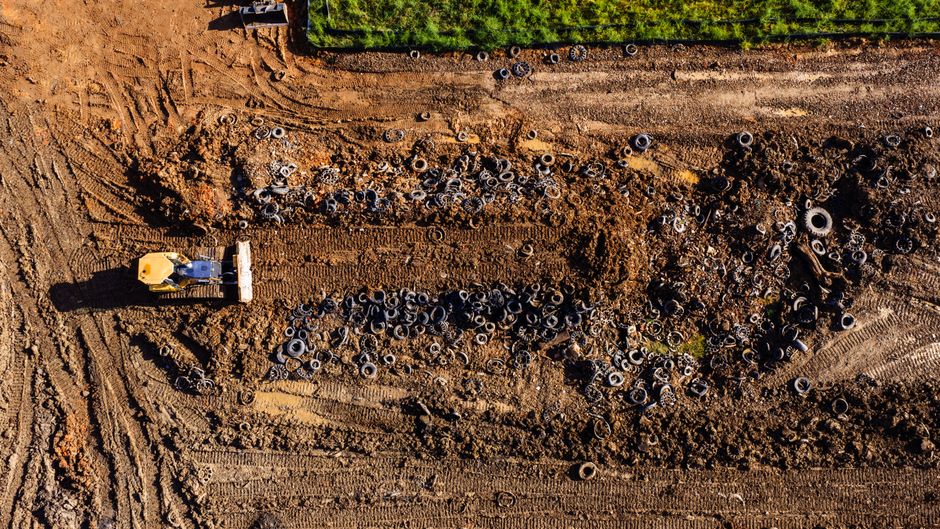 An aerial view of a garden undergoing landscaping work, featuring a large patch of freshly turned soil with visible tire tracks, bordered by a neatly trimmed green hedge. Scattered across the soil are