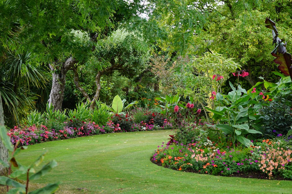 A vibrant garden scene featuring a well-maintained, lush green lawn bordered by colorful flower beds with pink, purple, orange, and white blooms. There are large leafy plants, and mature trees with te