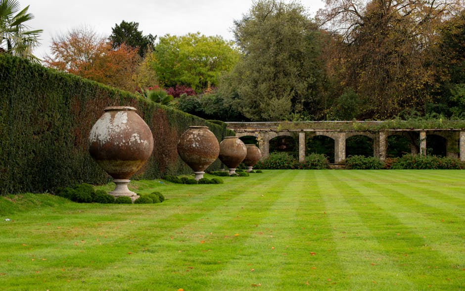 A vibrant garden scene featuring a well-maintained, lush green lawn bordered by colorful flower beds with pink, purple, orange, and white blooms. There are large leafy plants, and mature trees with te